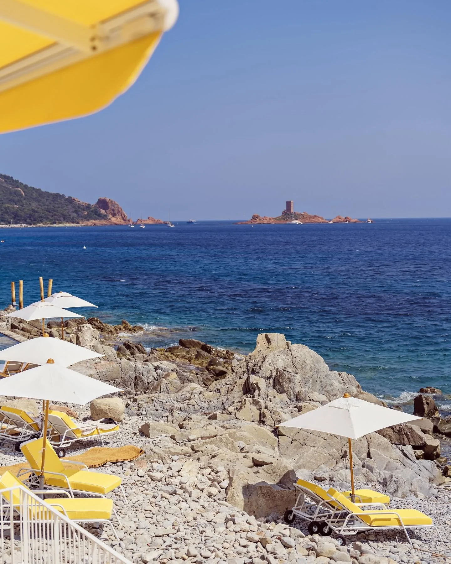 A rocky beach with yellow lounge chairs and white umbrellas, calm blue sea with boats, and a small island with a tower in the distance. A yellow and white umbrella in the top left corner partially covers the view.