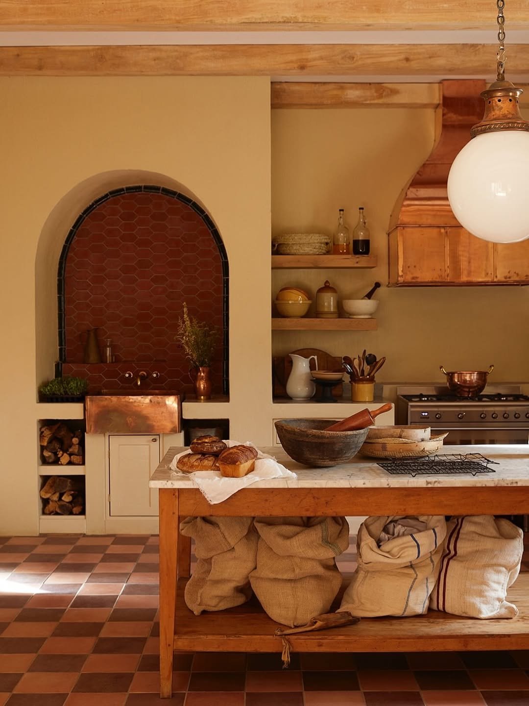 Kitchen with wooden elements, a marble-topped island with baked goods, sacks of grains underneath, vintage shelves with bowls, bottles, and kitchen tools, brick and wood accents, warm lighting.
