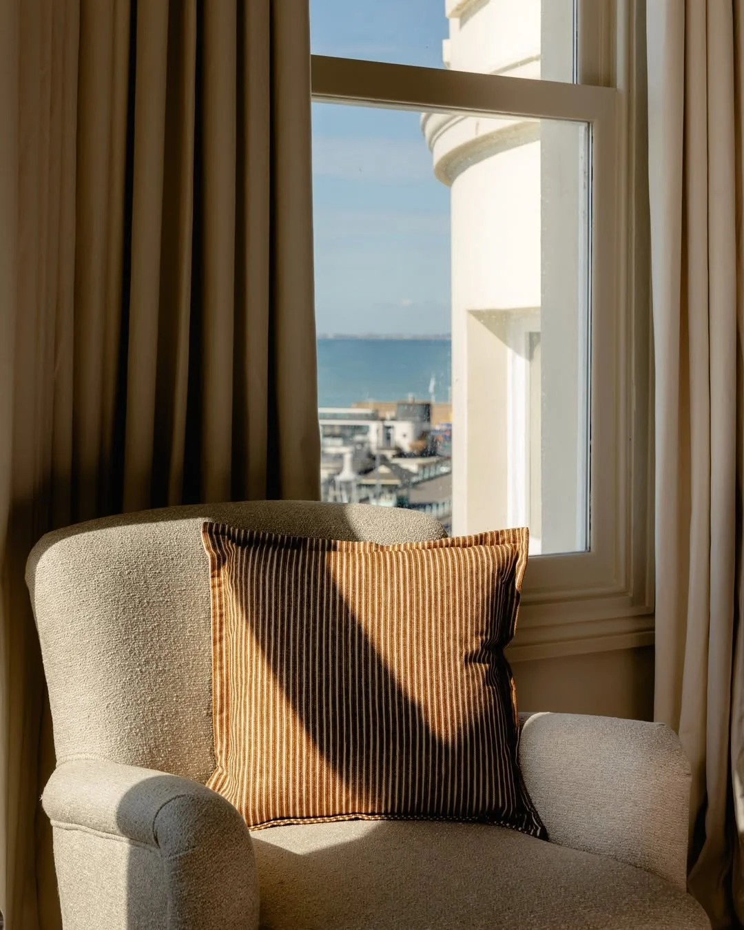 Sunlight coming through a window onto a beige armchair with a striped pillow, overlooking a cityscape and body of water outside.