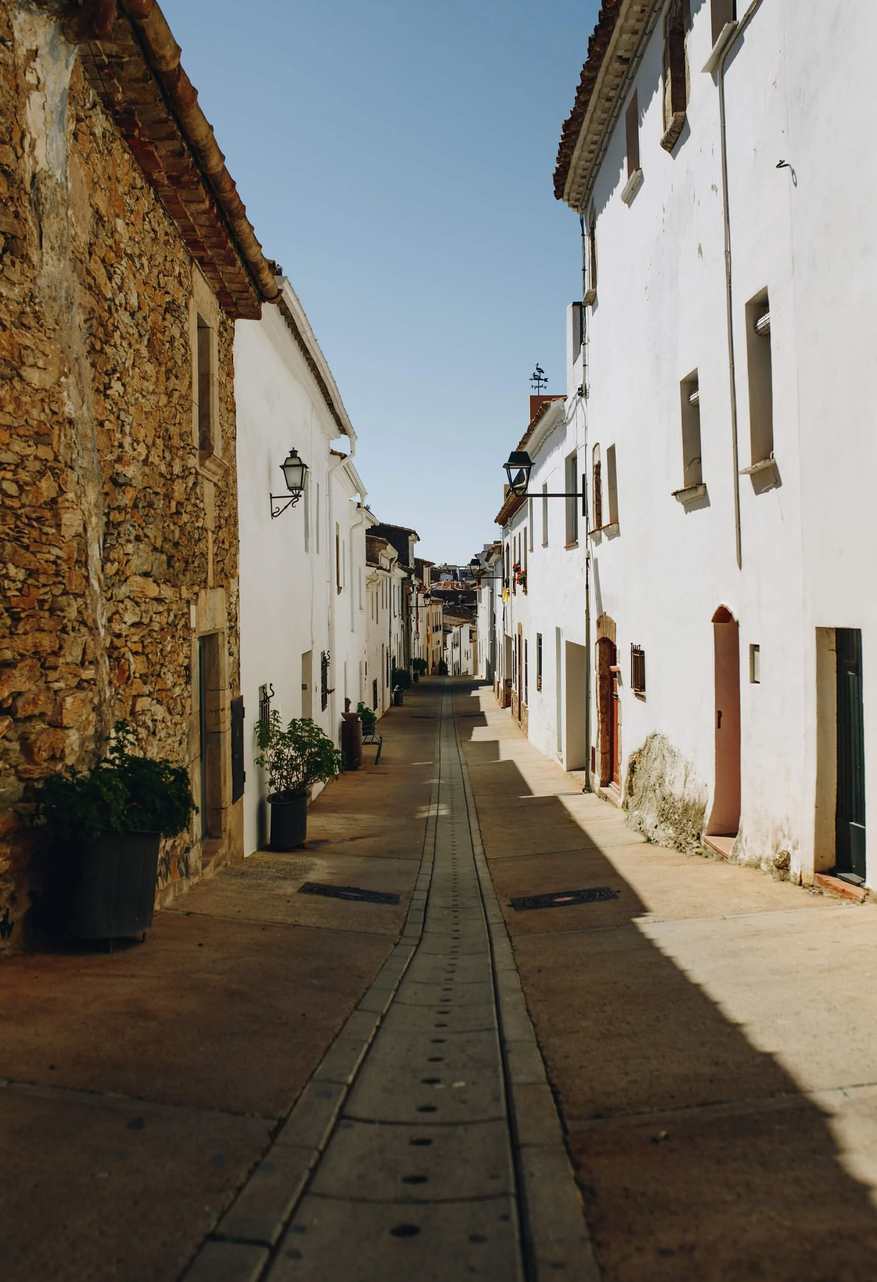 A narrow street lined with white buildings on both sides, with small windows and black wrought iron lamps, and a clear blue sky overhead.
