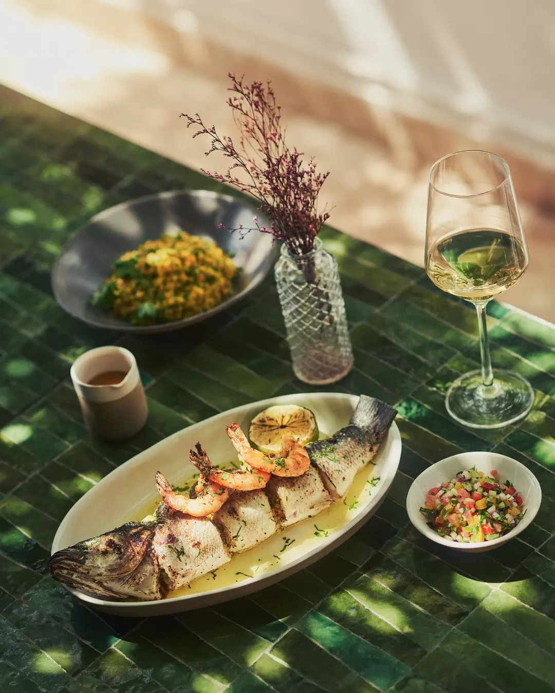 A table set with a whole grilled fish garnished with shrimp and lemon, a glass of white wine, and side dishes including salad, salsa, and a bowl of rice with vegetables. There is also a small cup of sauce, a vase with dried flowers, and a bowl of rice salad in the background.