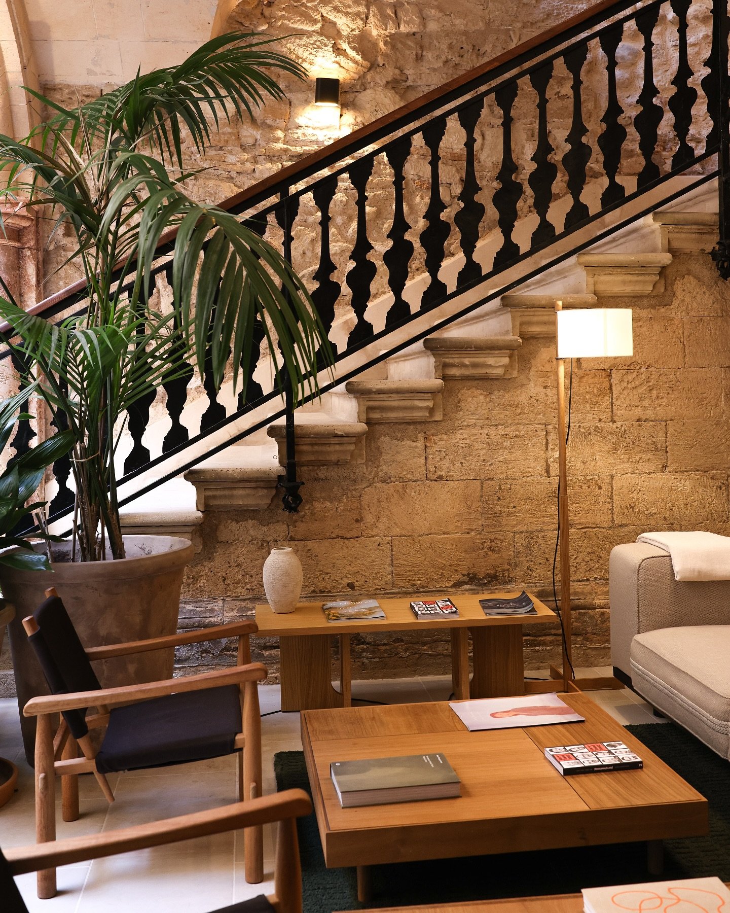 Interior of a stylish living room with a staircase, brick wall, potted plant, wooden furniture, magazines on the table, lamp, and a beige armchair.