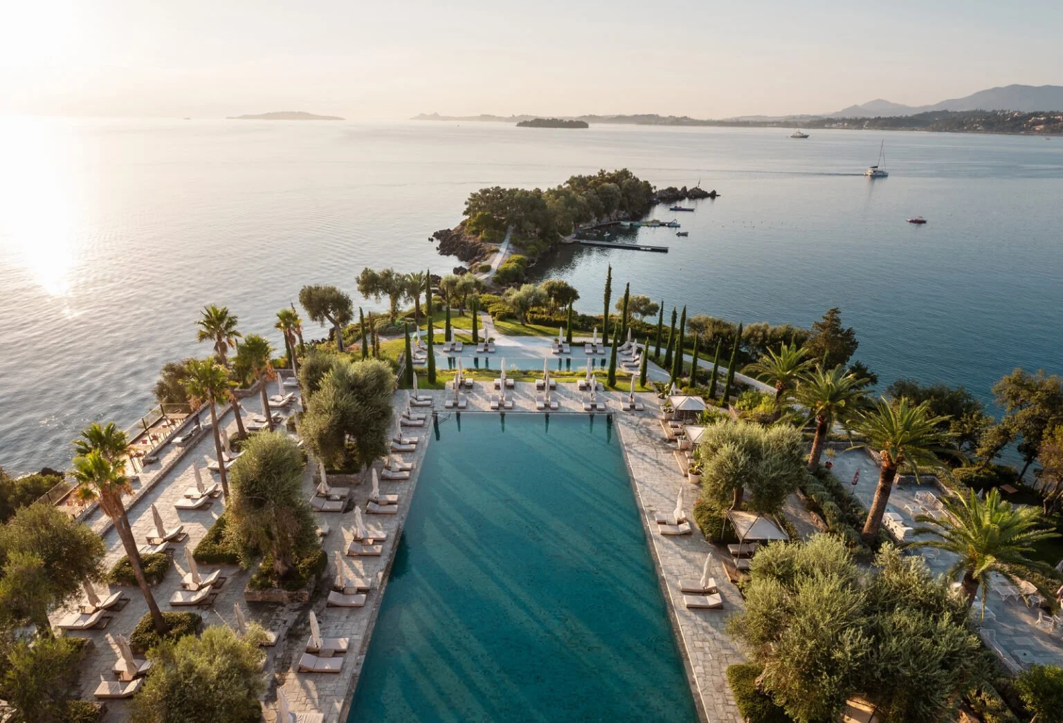 Aerial view of a luxurious seaside hotel with a large swimming pool lined with sun loungers and umbrellas, surrounded by palm trees and lush greenery, overlooking a calm bay with boats and islands in the distance.
