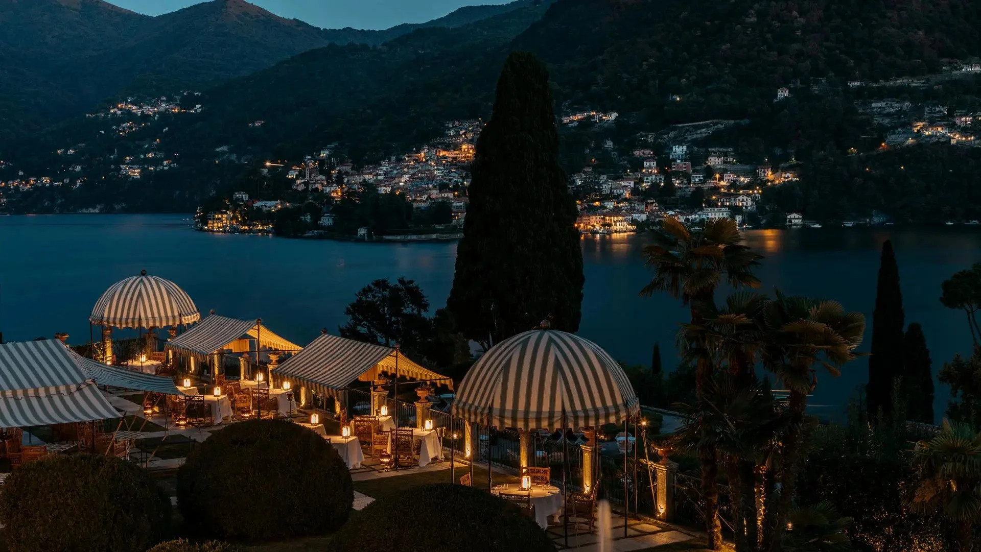 Outdoor dining area with striped umbrellas, tables, chairs, and lanterns overlooking a lake and hillside town at dusk.