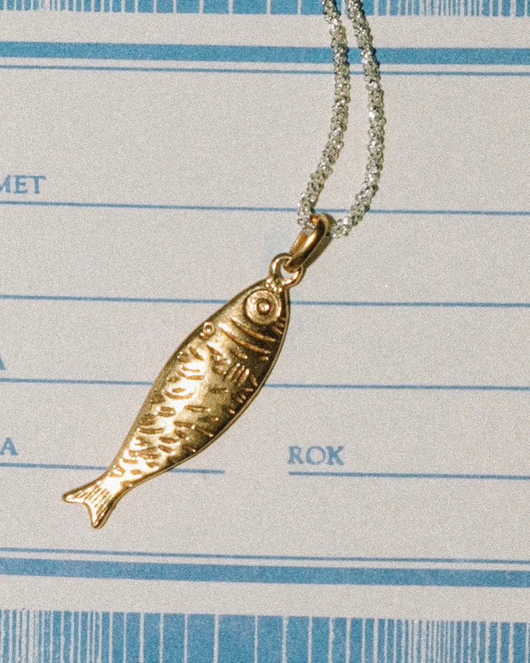 A silver fish-shaped pendant necklace resting on a piece of paper with blue lines and text.