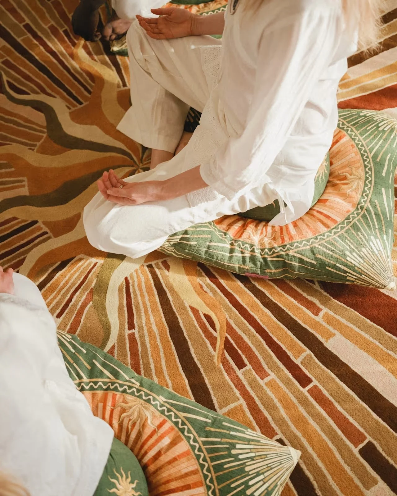 A person sitting cross-legged on a patterned cushion on the floor, wearing a white long-sleeved garment, with a decorative pillow under their legs, in a room with a colorful, abstract patterned carpet.