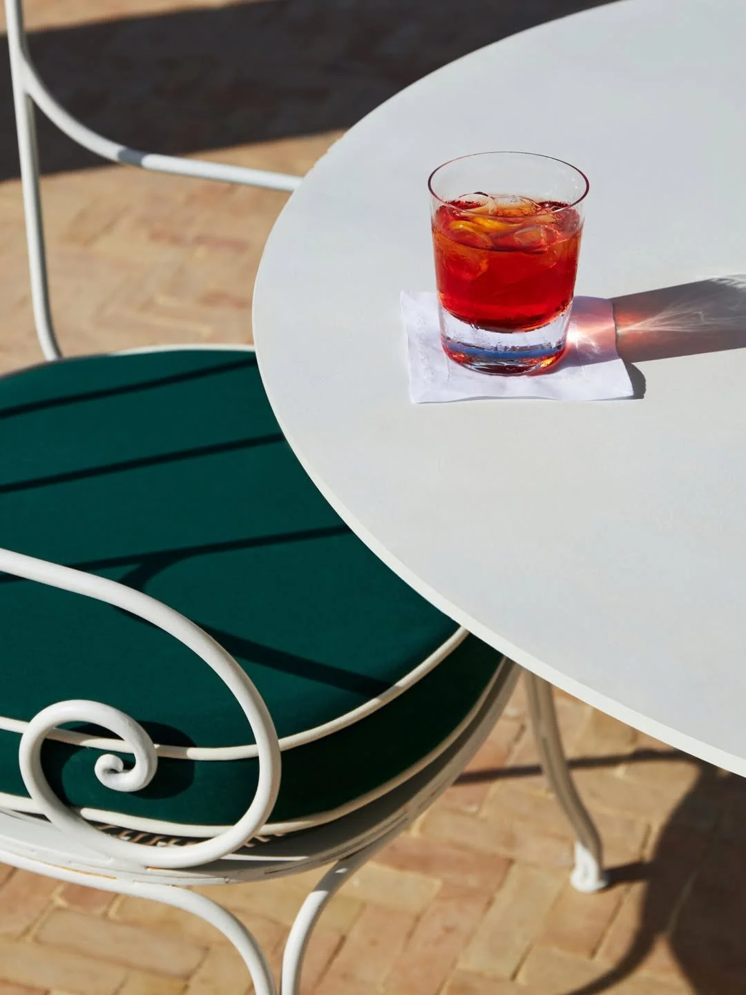 Glass of red iced beverage on a white round table with a white napkin. A green chair with a white metal frame is nearby, and the table is outdoors with a brick surface.