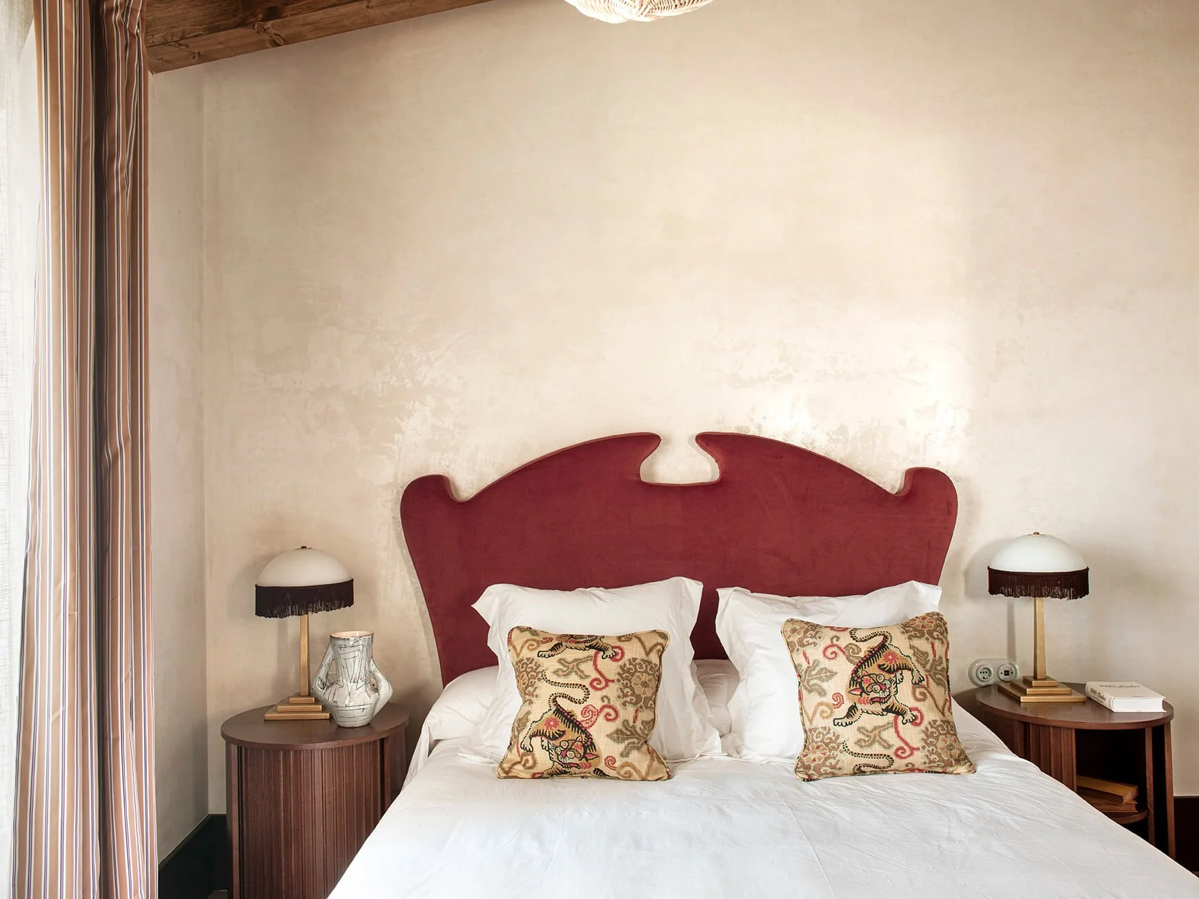 A hotel bedroom with a large bed, a red headboard, four embroidered pillows with lion designs, two bedside tables with lamps, a decorative glass vase, a book, and striped curtains.