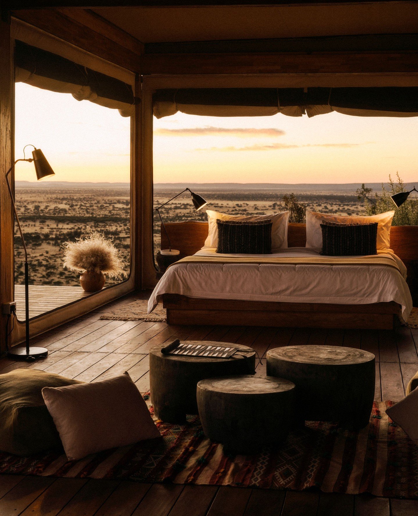 Interior of a cozy bedroom with large windows showing a desert landscape at sunset, featuring a bed with white and black pillows, bedside lamps, a potted plant, and wooden stools.