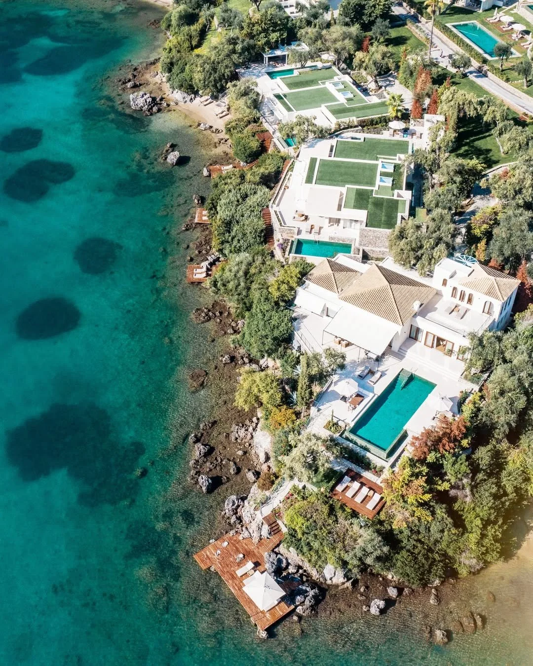 Aerial view of a luxurious beachfront estate with multiple pools, surrounded by lush greenery and rocky shoreline, with a wooden dock extending into the clear blue water.