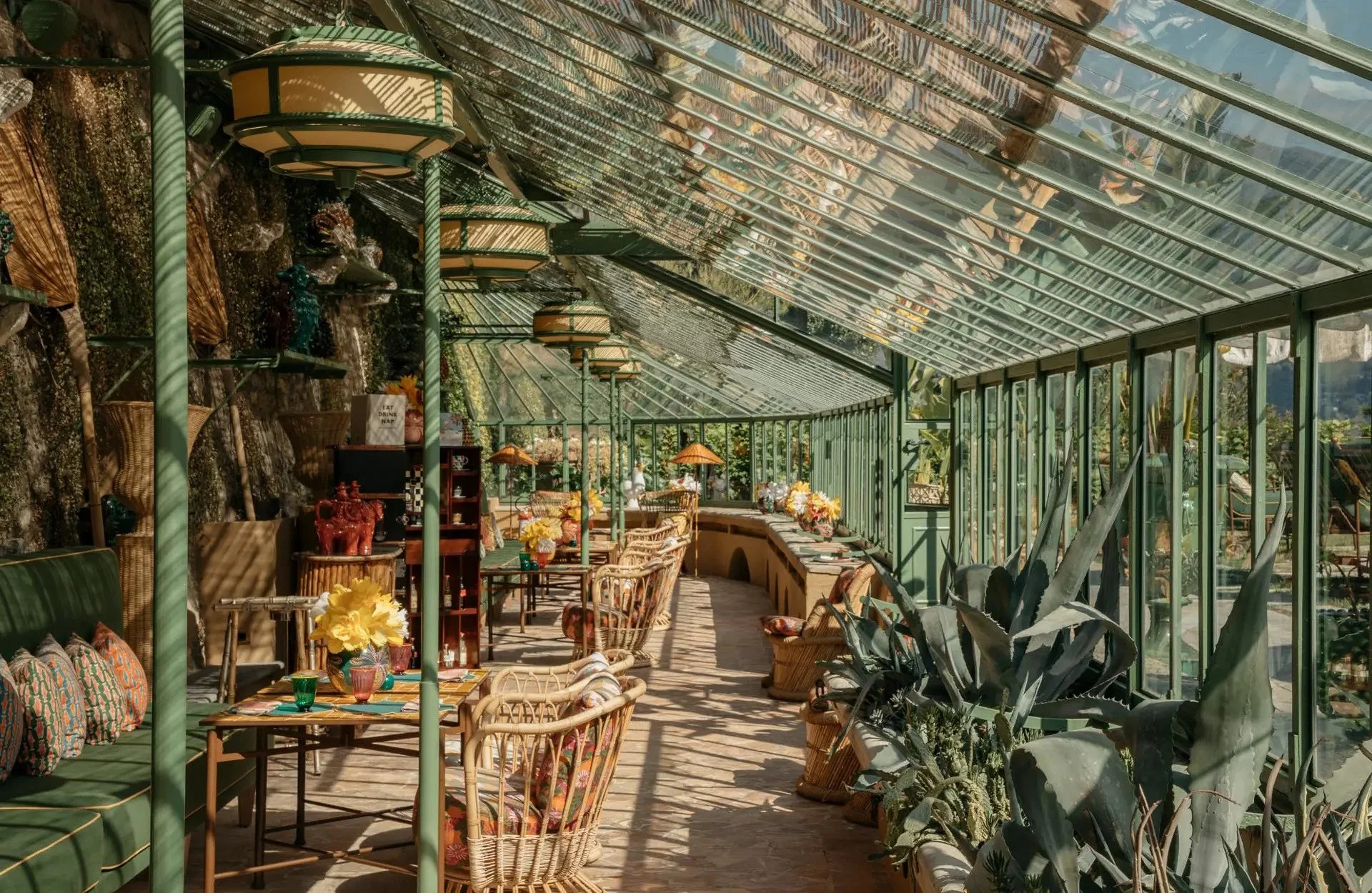Indoor greenhouse dining area with wicker chairs, glass walls, potted plants, and decorative lamps.