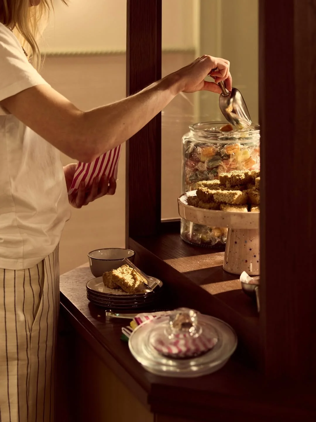Person serving fruit candy from a glass jar onto a plate of cookies in a nostalgic kitchen setting.