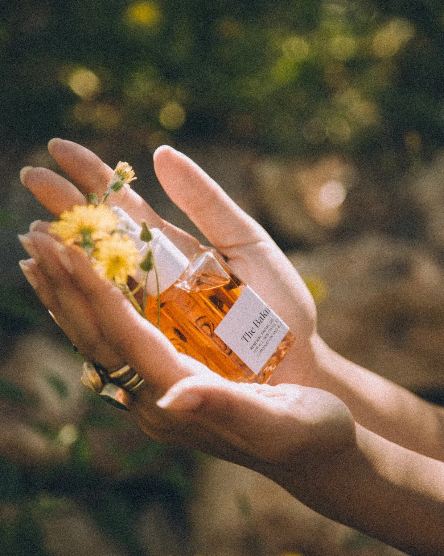 A person's hand holding a bottle of amber-colored perfume with a white label and yellow flowers attached to it.