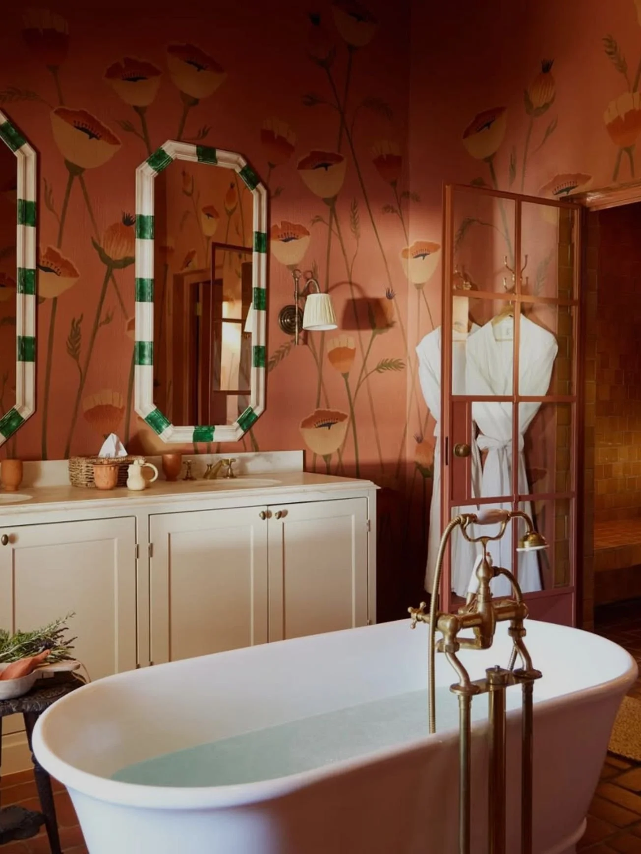 A bathroom with a vintage style, featuring a white freestanding bathtub filled with water and a brass faucet. The wall has pink floral wallpaper, and above the white vanity with a sink, there are two mirrors with green and white striped frames. A person in a white robe is visible through a partial glass-paneled door, and various decorative items are on the vanity. The bathroom has warm lighting.