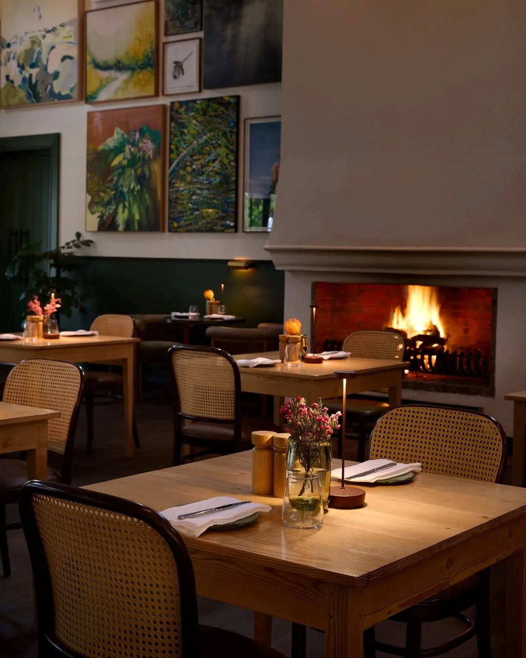 A cozy restaurant interior with wooden tables set with white napkins, glassware, and small flower arrangements. A fireplace with a fire adds warmth to the room. Artwork adorns the wall above the tables, and soft lighting creates an inviting atmosphere.