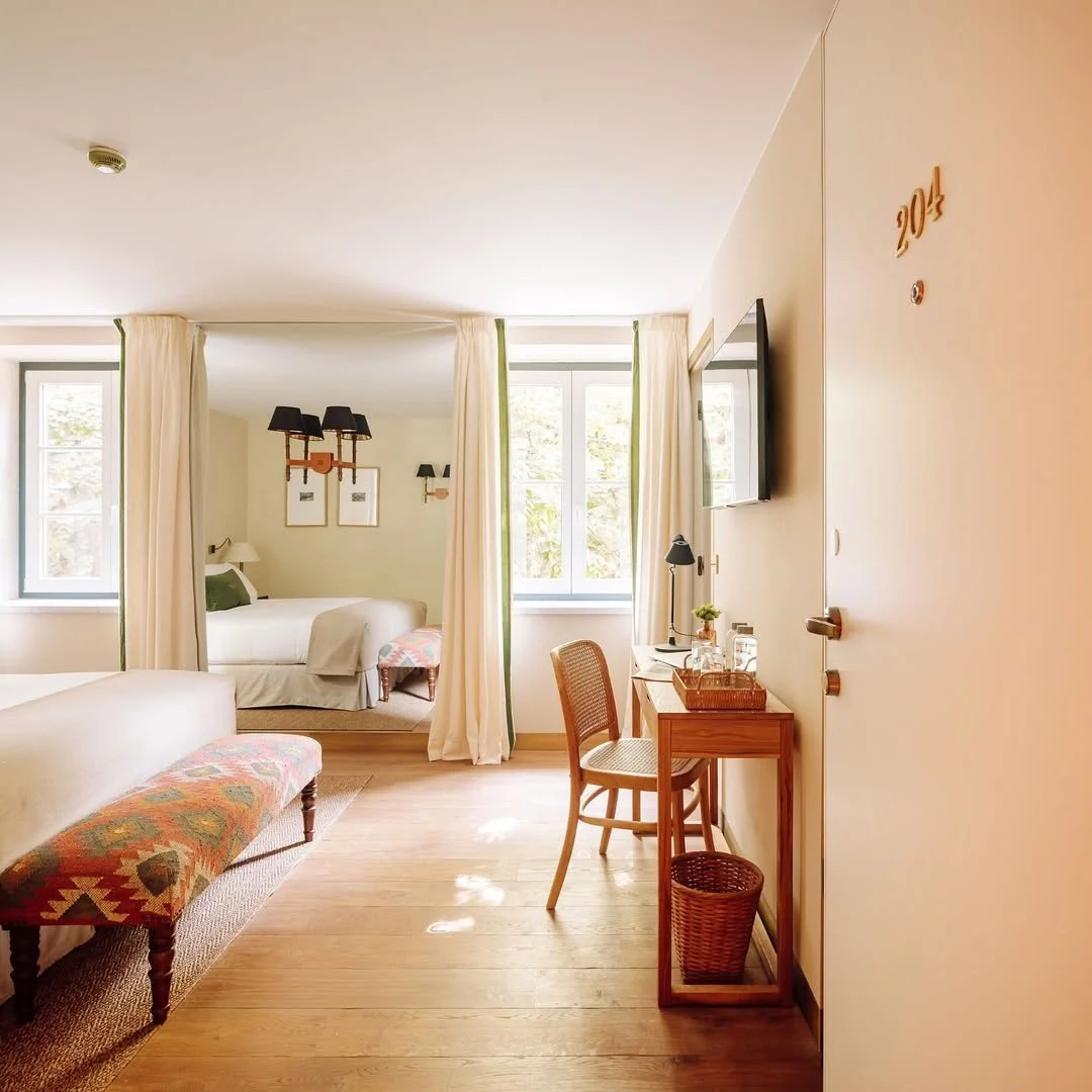A cozy hotel room with two beds, a small desk with a chair, a wall-mounted TV, and large windows with cream-colored curtains, decorated in a warm, minimalist style.