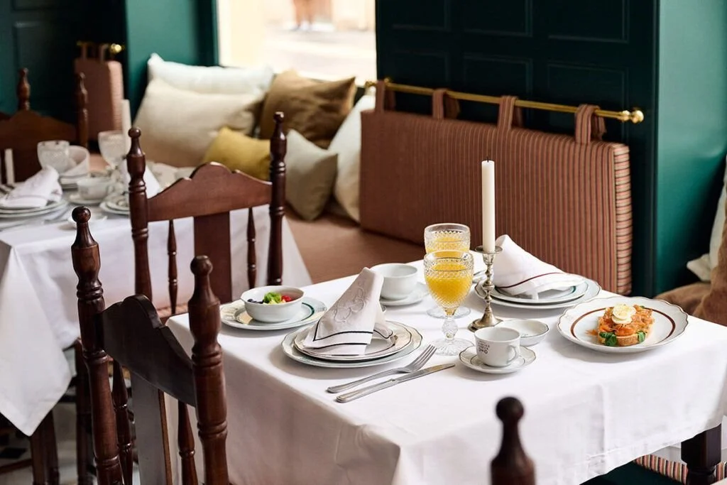 A dining table set for breakfast with white tablecloth, plates, bowls, glasses of orange juice, silverware, a white candle, a coffee cup, and a plate with a meal. In the background, there are chairs, pillows, and a cozy seating area with a bench and cushions.