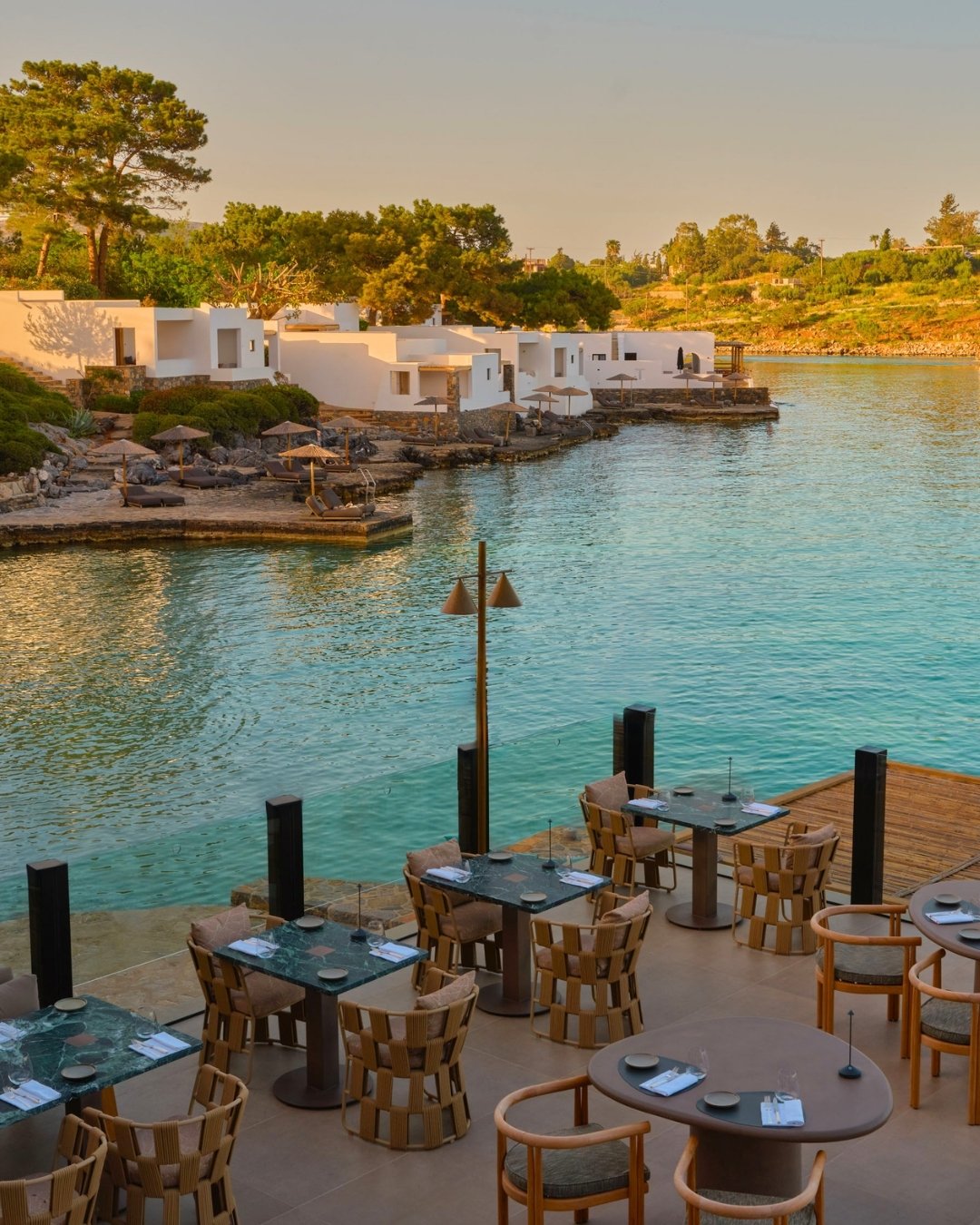 An outdoor restaurant with tables and chairs set near a body of water, with a shoreline of white buildings and trees in the background.