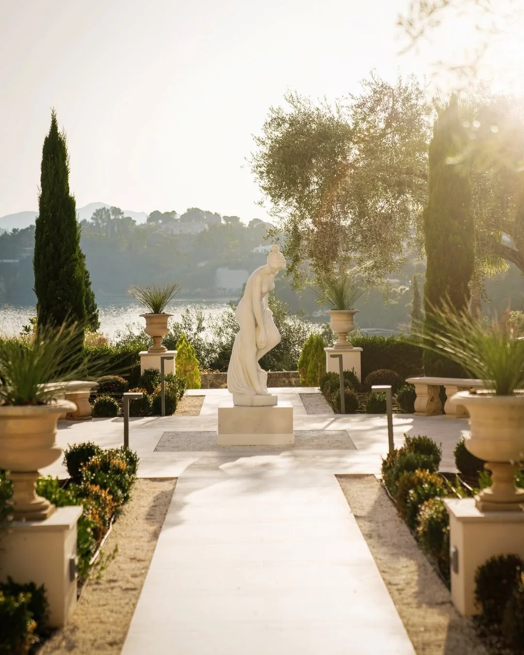A marble statue of a woman on a pedestal in a landscaped garden with potted plants, tall cypress trees, and a view of a lake or river with hills in the background, bathed in warm sunlight.