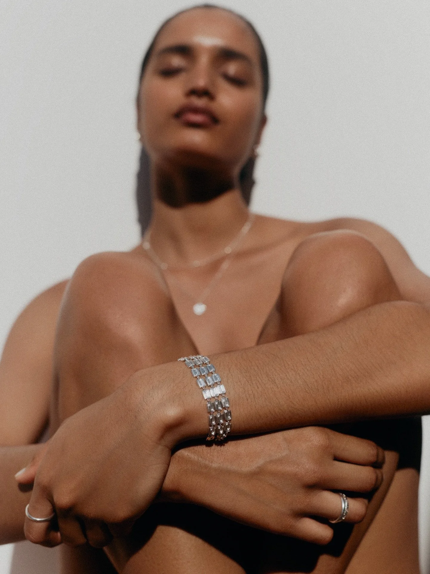 A woman with dark skin and short black hair is posing with her arms crossed over her chest. She is wearing multiple jewelry pieces, including a silver bracelet, rings on her fingers, and a necklace. The background is plain white.