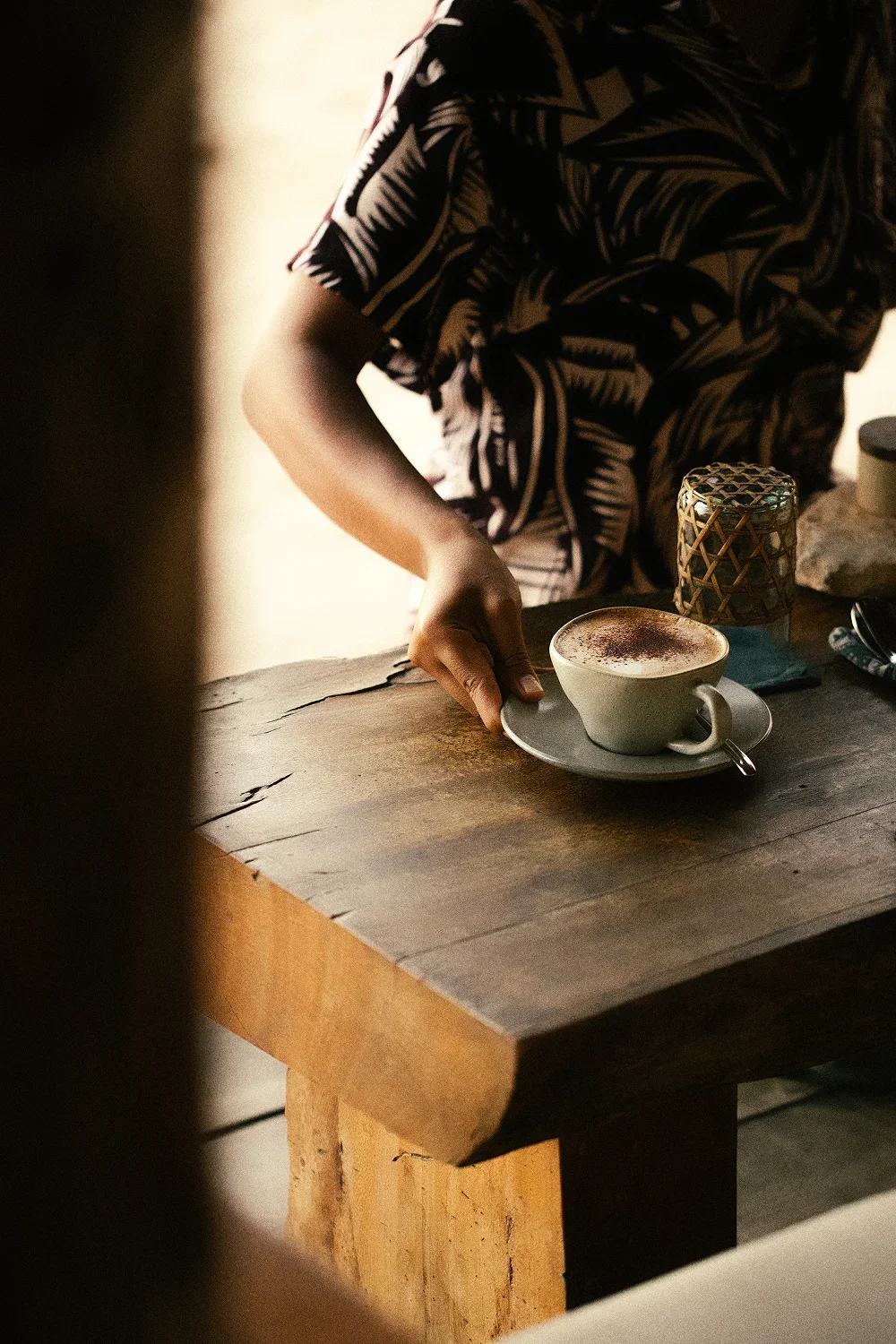 Person placing a cup of coffee or cappuccino on a gray saucer on a rustic wooden table.