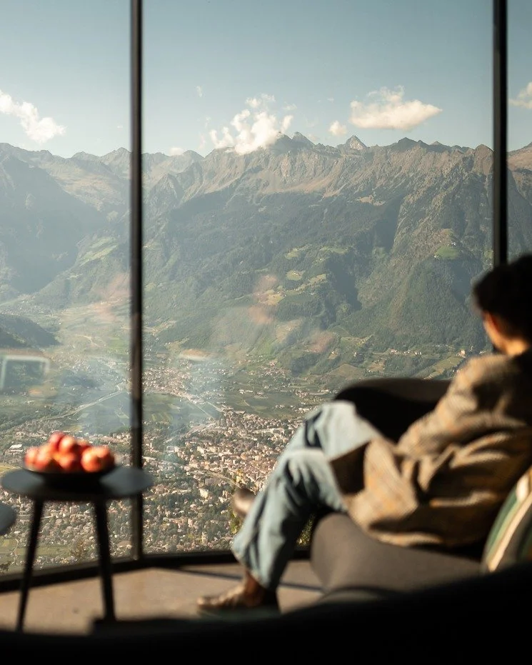 Person sitting on a couch looking out a large window at mountainous landscape and a town below, with a small table holding a bowl of fruit nearby.
