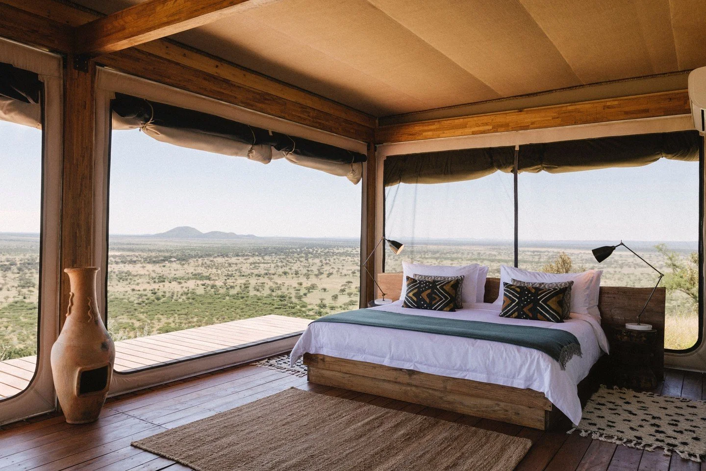A bedroom with a large bed, white bedding, and black and gold pillows. The room has wooden beams and floors, with large windows offering a view of a flat landscape and distant mountains.