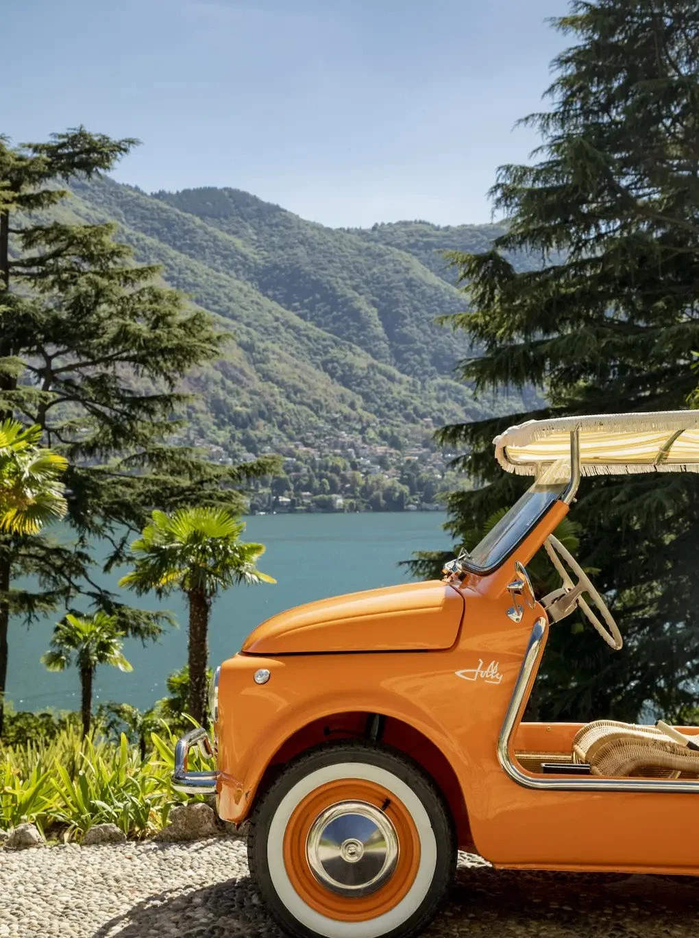 Orange vintage golf cart with a white canopy, parked on pebbled ground near green plants and tall trees, with a lake and green mountains in the background under a clear blue sky.