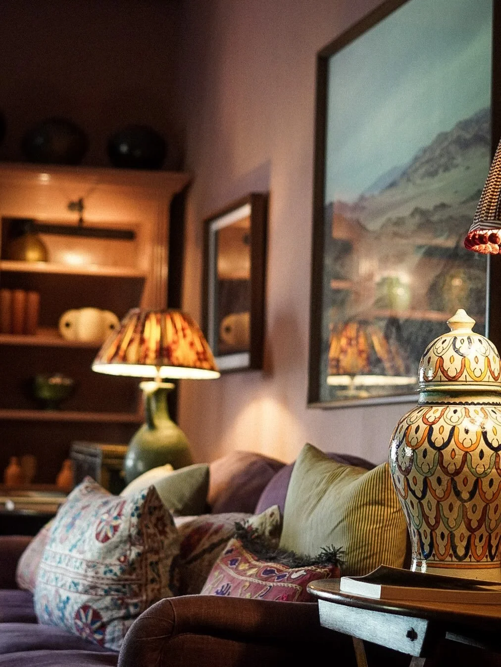 Cozy living room with a patterned ceramic lamp, colorful pillows on a purple couch, a picture on the wall, and a bookshelf in the background.