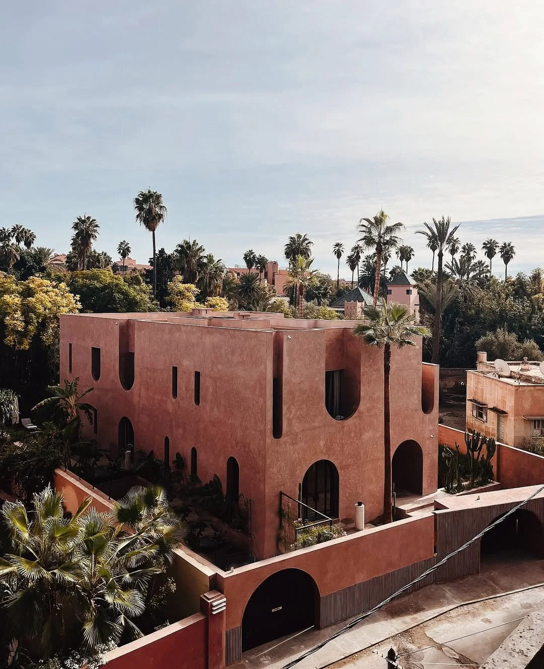 A modern pink building with curved and rectangular windows, surrounded by palm trees and lush greenery, under a clear sky.