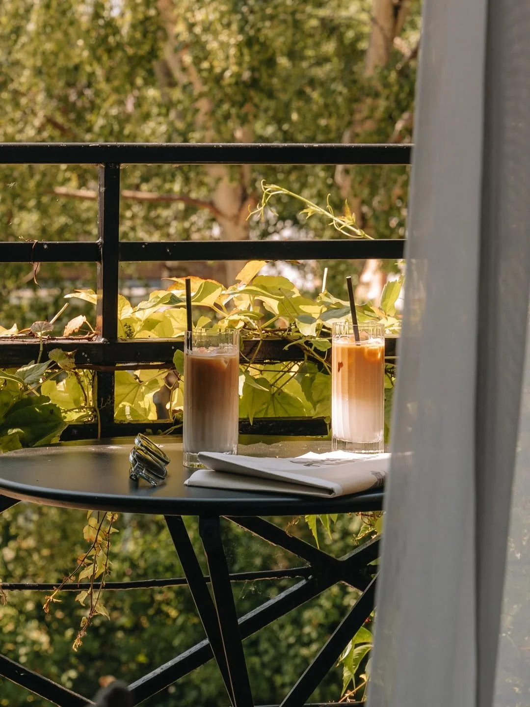 Two glasses of iced coffee on a balcony table, with a napkin and keys, surrounded by green leafy plants and trees outside.