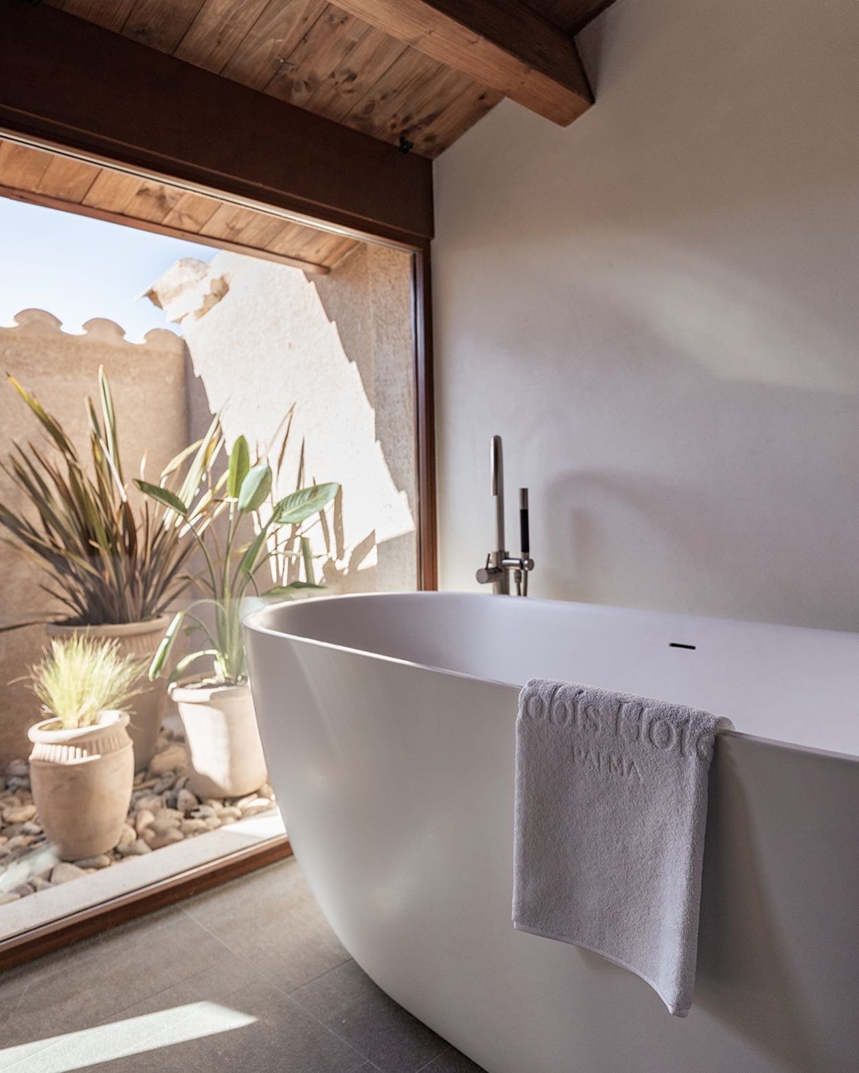 Free-standing white bathtub with a towel hanging on its side, positioned next to a large window revealing potted plants outside, in a bathroom with a wooden ceiling.