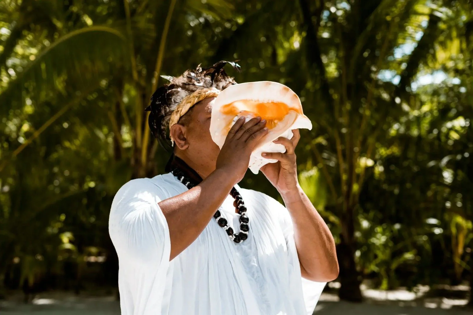 A person with traditional attire holding a large shell to their face, standing outdoors among trees and greenery.