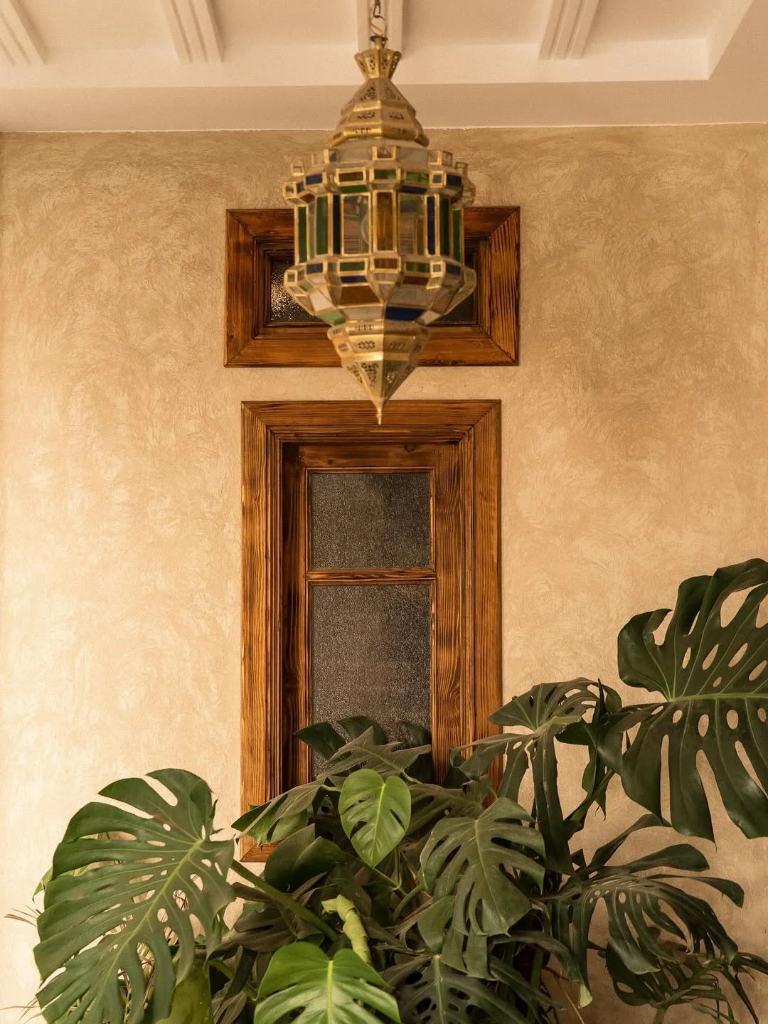 Decorative hanging lantern above a wooden framed window and large Monstera plant in front of a beige textured wall.