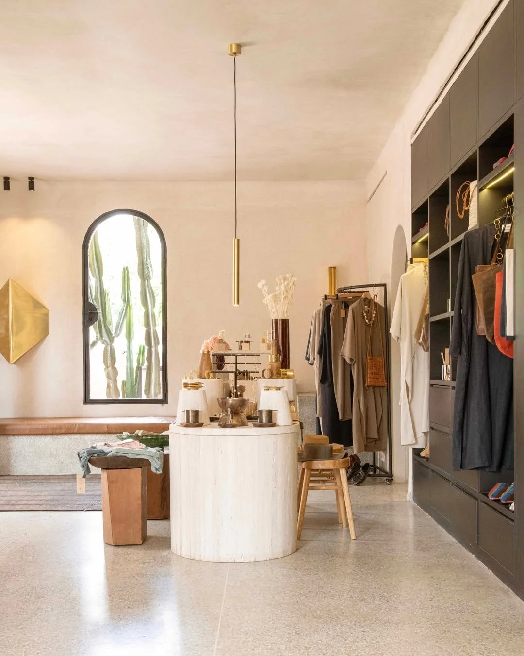 Interior of a boutique clothing store with a black shelving unit displaying clothes, a clothing rack, a round white display table with decor, stools, a window with cactus plants outside, and modern lighting.