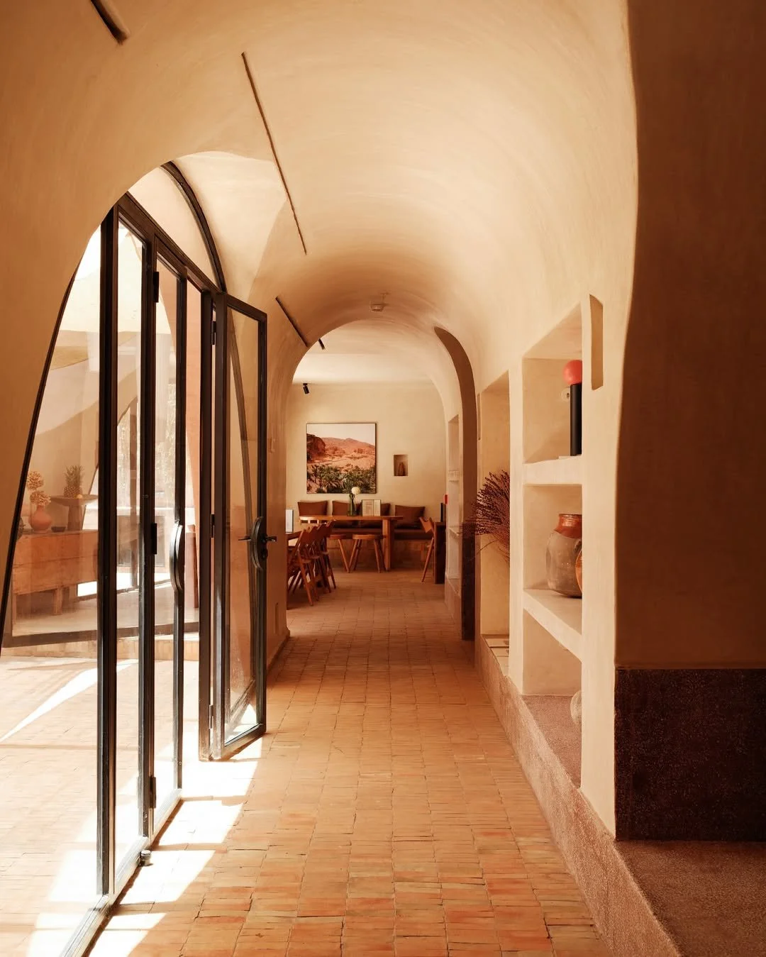 A hallway with a curved ceiling, brick flooring, and built-in wall shelves containing pottery. Glass doors on the left lead to another room, and at the end, there is a dining area with a table, chairs, and a landscape painting on the wall.
