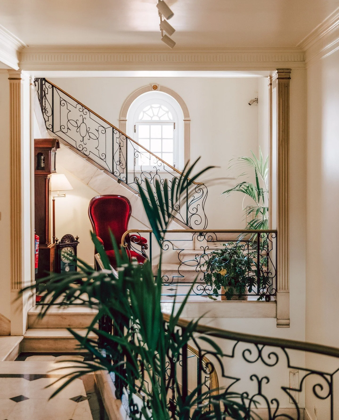 Interior view of a house with a staircase featuring ornate black iron railing, a white marble steps, a red velvet armchair, a tall wooden cabinet, plants, and a window with decorative trim in the background.