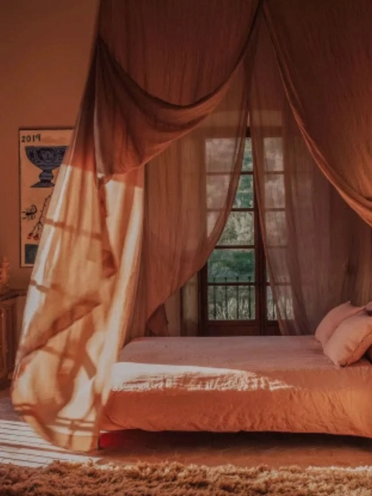 A cozy bedroom with a four-poster bed draped with sheer orange curtains. A large window behind the bed reveals a view of trees and greenery. The room has wooden walls and a patterned rug on the floor.