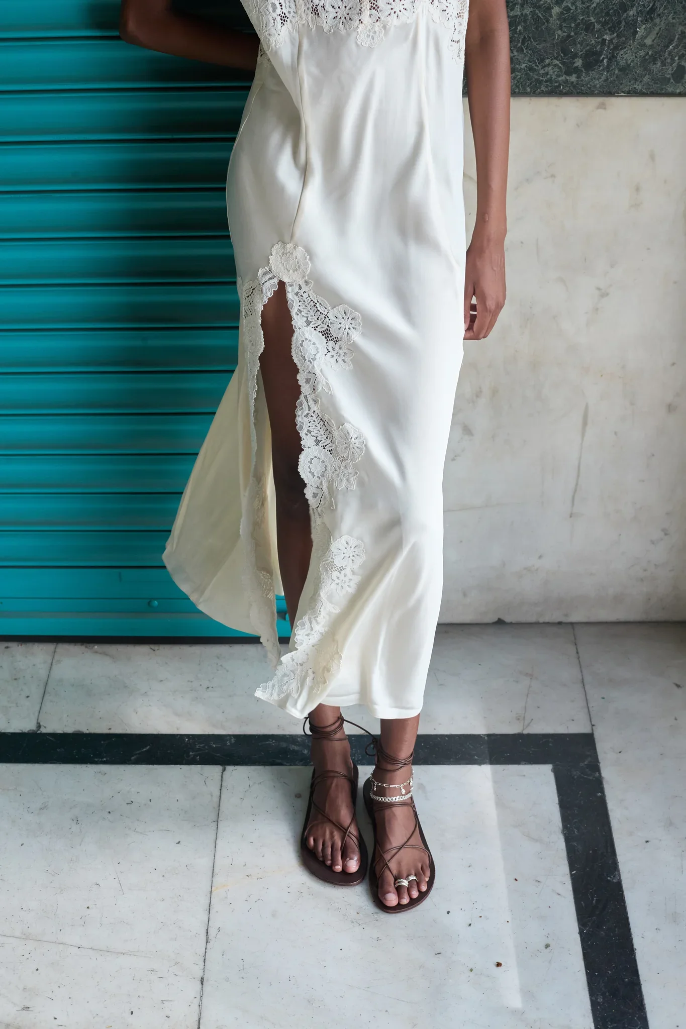 A woman wearing a white dress with lace details, standing on a tiled floor with black and white stripes, wearing brown lace-up sandals and ankle jewelry.