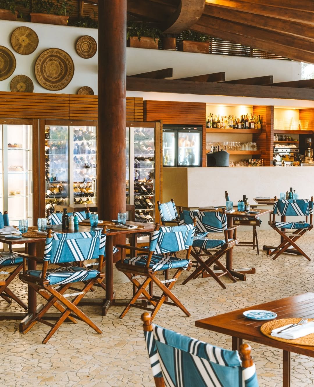 Interior of a restaurant with wooden furniture, blue-striped chairs, glassware on tables, a wine cooler, and a well-stocked bar in the background.
