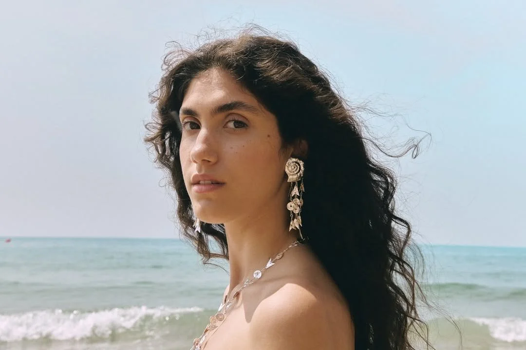 A woman with dark, curly hair at the beach, wearing large floral earrings and a necklace, with ocean waves in the background.