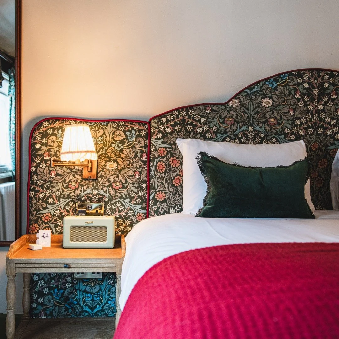 A hotel bedroom with a floral-patterned headboard, a white pillow and a dark green pillow on a bed with white sheets and a red blanket. A bedside table holds a vintage radio, an alarm clock, and a small box. A wall-mounted lamp is turned on.