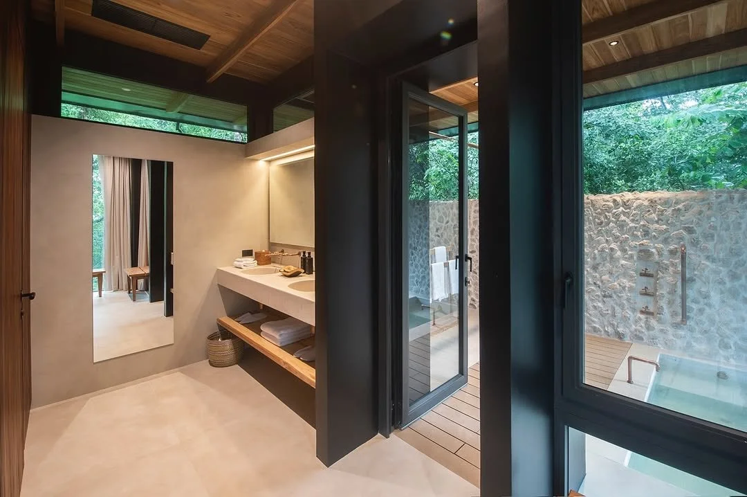 Modern bathroom with a large mirror, a countertop with towels and toiletries, an open door leading to outdoor shower area with stone wall and greenery.
