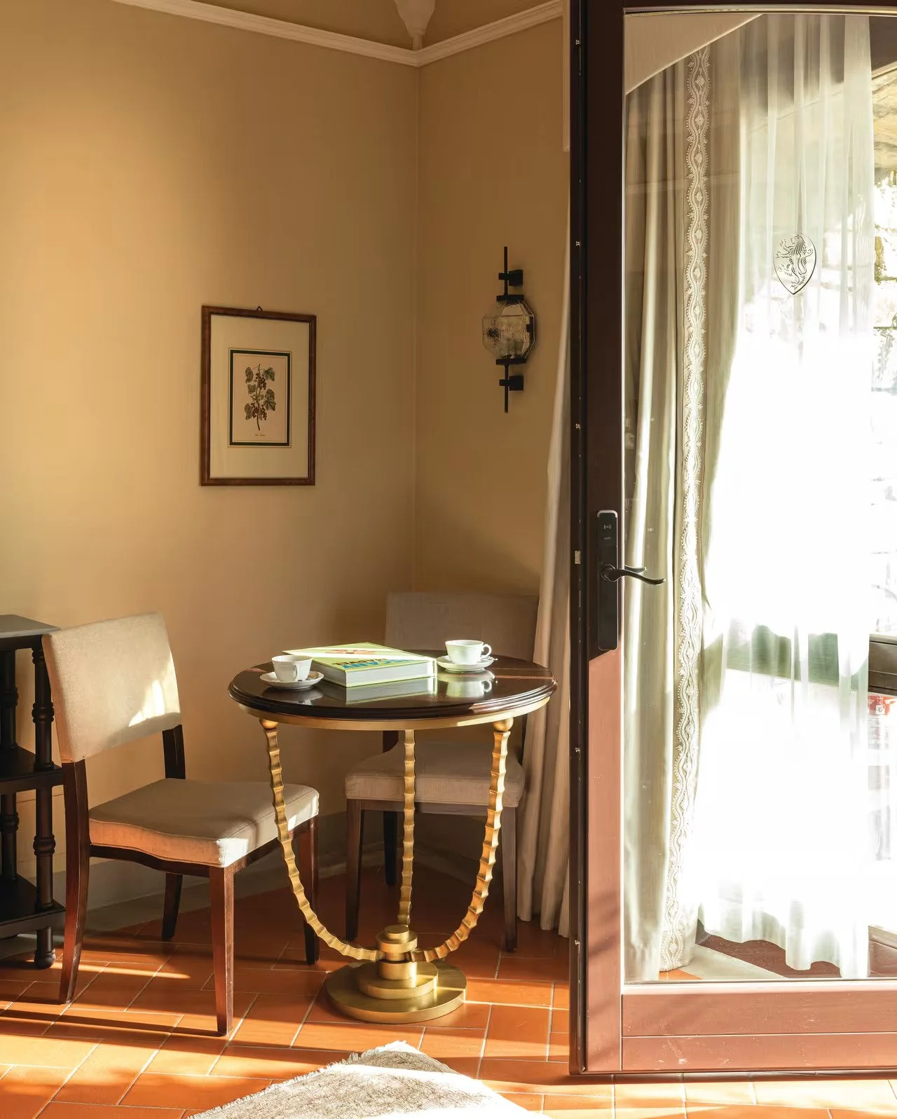 A cozy corner in a room with beige walls, a framed botanical print, and a black wall-mounted candle holder. There is a small round wooden table with golden legs, set with a book and two white cups and saucers. An open sliding glass door with sheer curtains leads to a sunlit balcony.