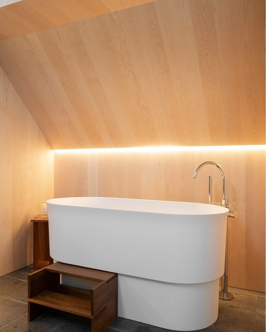 Modern white bathtub with a wooden step and small side table, wall-mounted faucet, wood-paneled walls with LED lighting, in a minimalist bathroom.