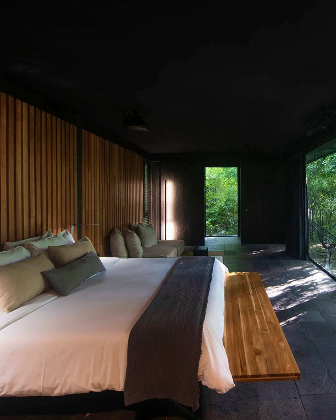 A modern bedroom with a large bed, multiple pillows, a wooden bench at the foot of the bed, dark walls, a wooden accent wall, and a glass door leading to a balcony with greenery outside.