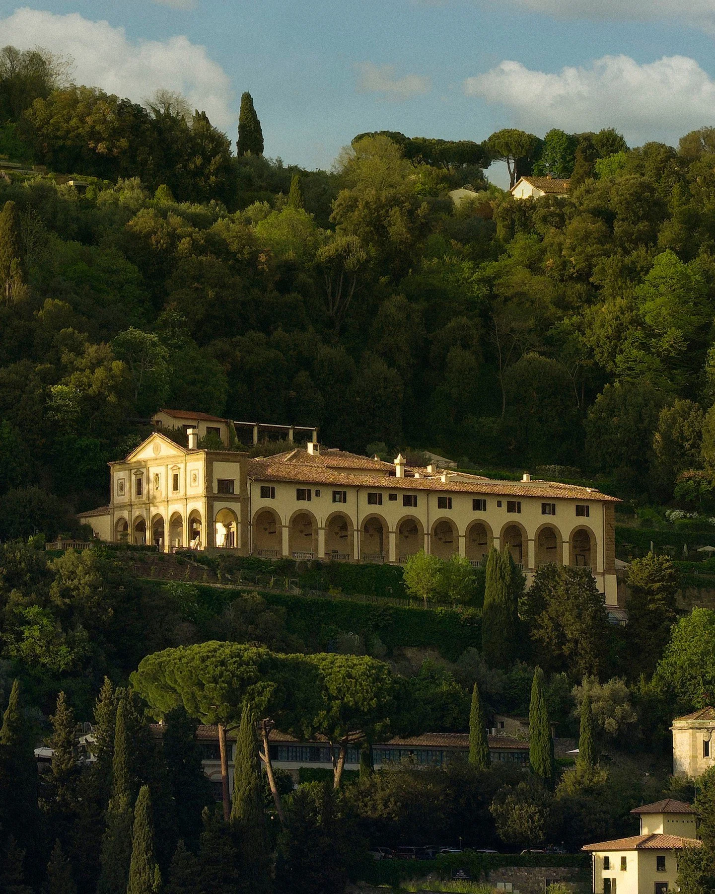 A large, luxurious estate with a white Mediterranean-style villa featuring arches and a tiled roof, nestled on a lush hillside covered with green trees and foliage.