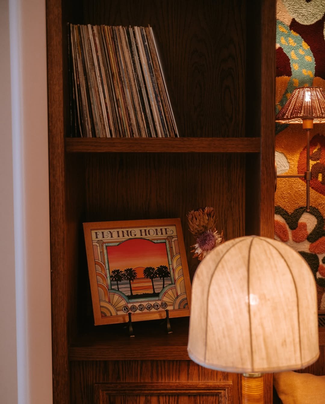 A wooden bookshelf with a vinyl record collection on top shelf, a framed picture titled 'Flying Home Summer' with sunset and palm trees on the bottom shelf, and a beige desk lamp with a woven shade in front.