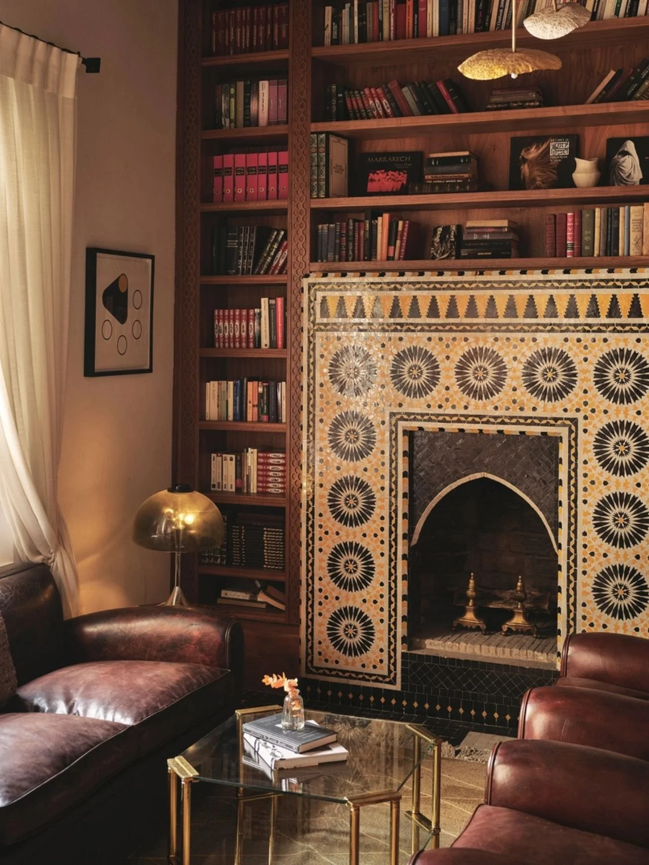 A cozy living room with a fireplace decorated with intricate Moroccan-style tile work, surrounded by wooden bookshelves filled with books and decorative items. There are leather armchairs, a glass coffee table with books and flowers, and a brass floor lamp. The room has a warm, inviting atmosphere with soft lighting and curtains on the window.