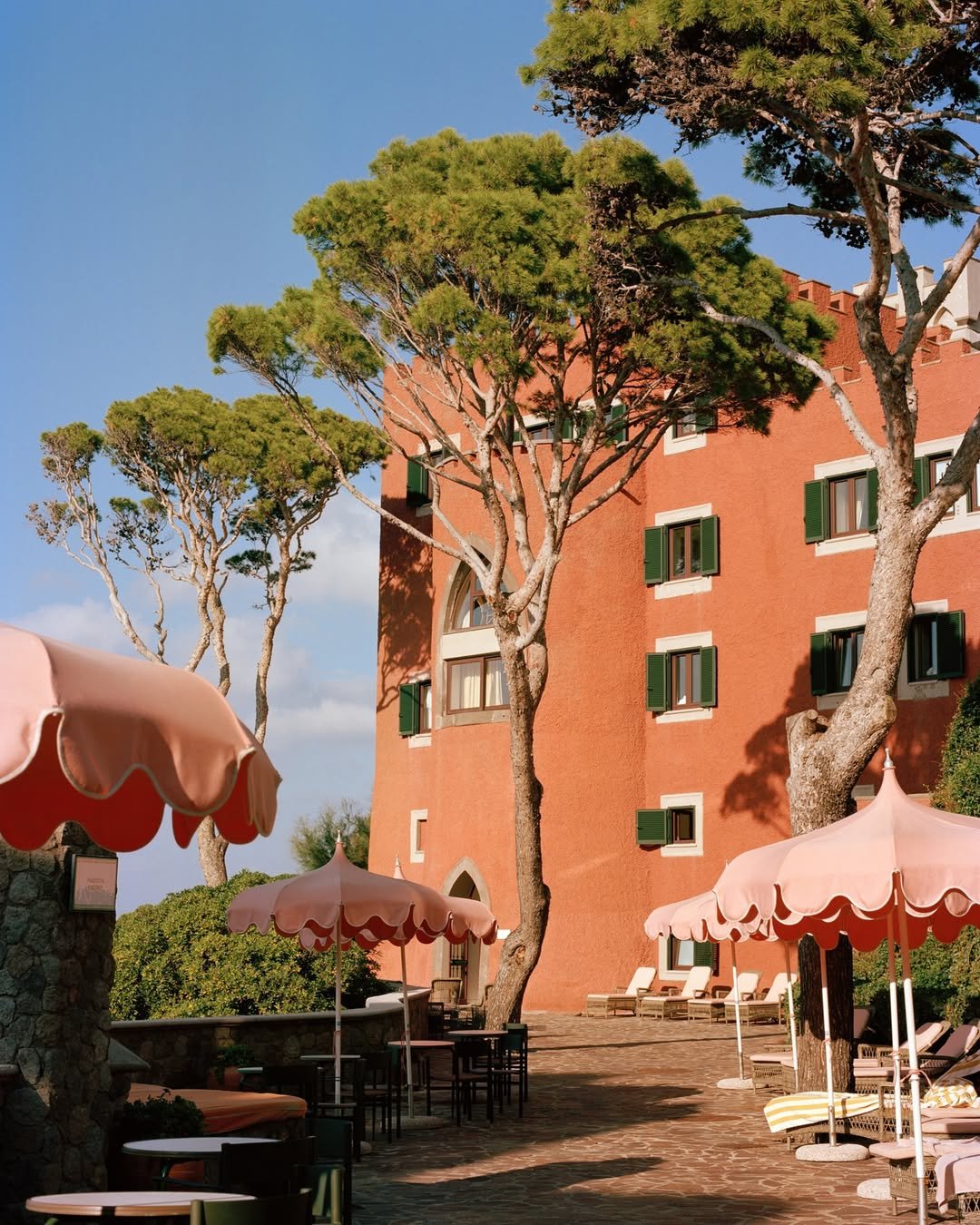A courtyard with pink umbrellas, lounge chairs, and a tall red building with green shutters, surrounded by trees under a clear blue sky.